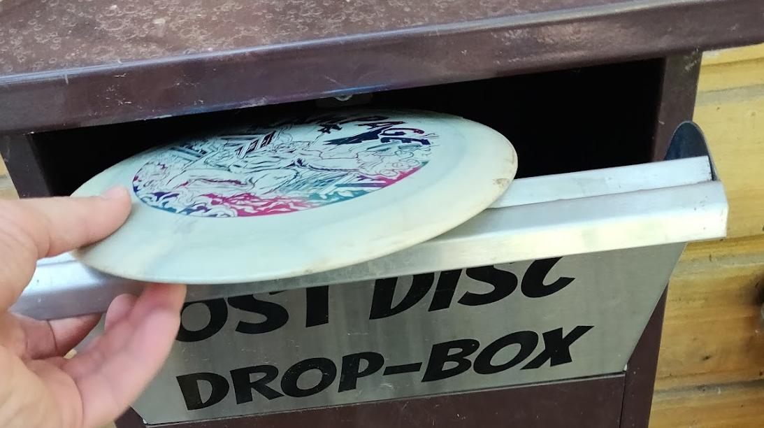 A disc golf disc being placed into a lost disc drop box.
